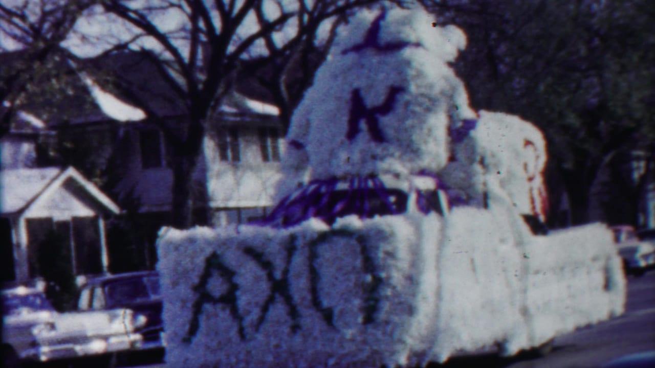 1958 Cheyenne County, Kansas frame showing the flower-covered float closer to camera, with AXO letters, a large K, houses, and parked cars.