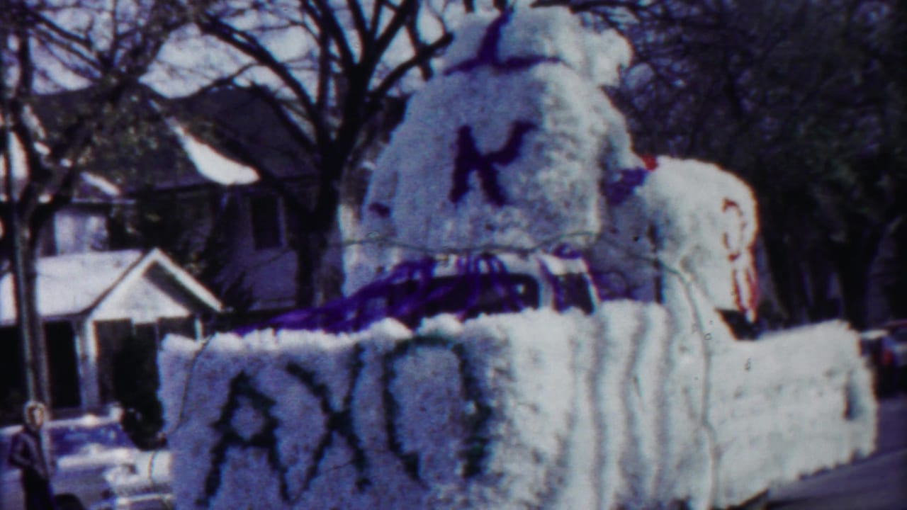 Home-movie frame from 1958 Kansas showing the decorated white float moving along a residential street, with a small spectator figure near the curb.
