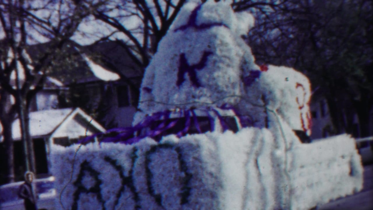 Decorated parade float with white floral covering, purple trim, and a large figure on a residential street.
