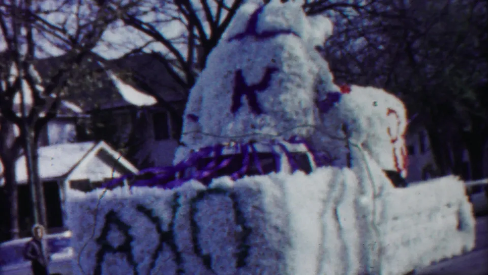 1958 Cheyenne County, Kansas home-movie frame showing a white floral parade float with purple trim passing houses and bare trees.
