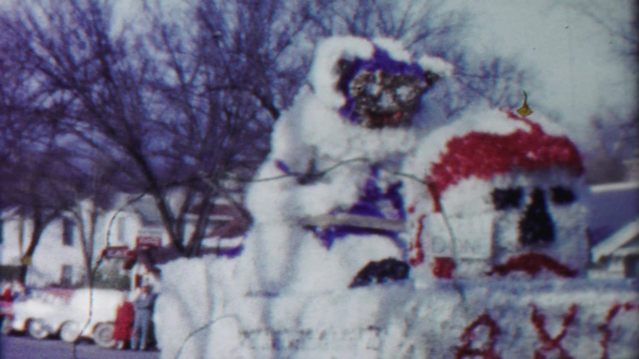 A white float in a Cheyenne County, Kansas parade shows a large decorated figure, red facial accents, bare trees, and people along the route.