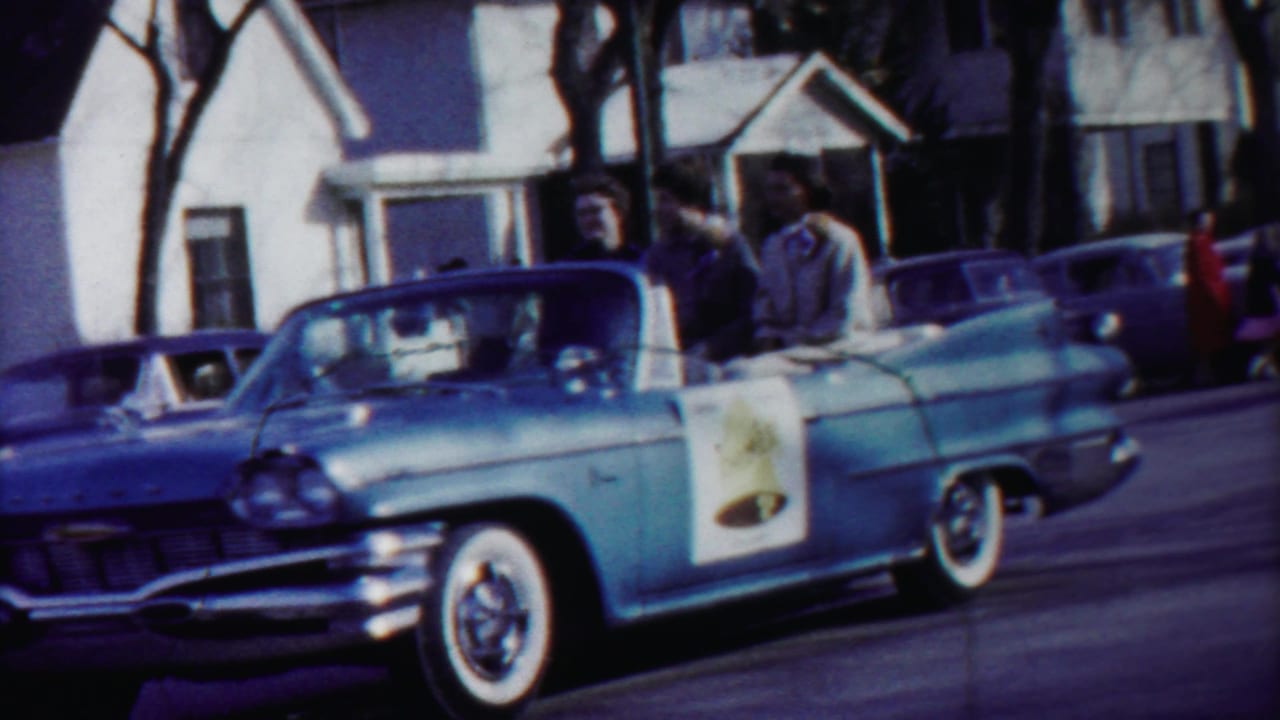 The blue convertible continues past the camera on a residential parade route, with passengers visible and houses behind it.