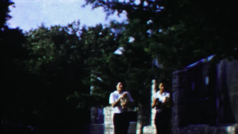 Two women in light blouses and dark skirts walk toward the camera in 1958 rural Kansas near trees and a stone roadside feature.