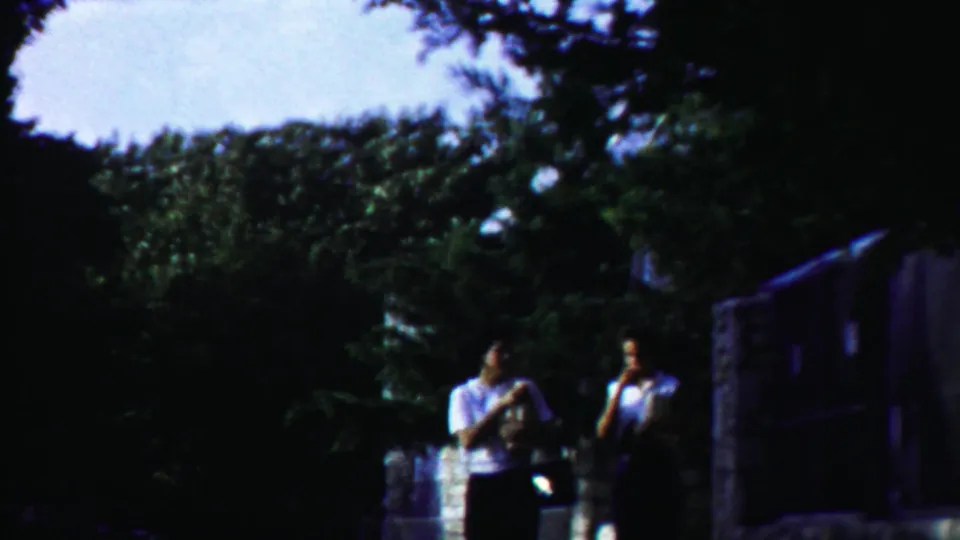 Two women continue along the rural path in a 1958 Kansas home movie, with trees, stonework, and a dark roadside structure nearby.