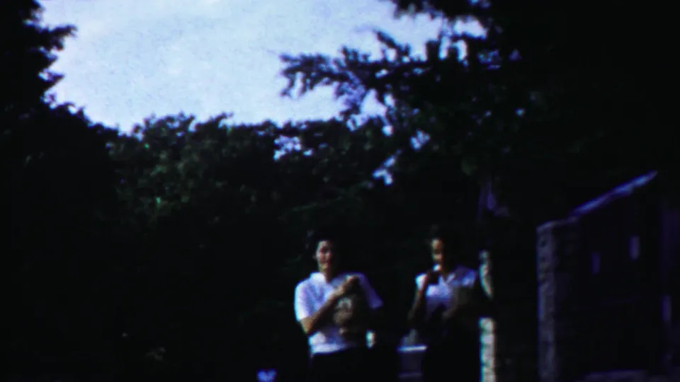 The pair walks closer to camera in a leafy Cheyenne County, Kansas setting, with one holding a small object against her chest.