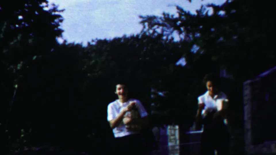 Two young women in 1958 Kansas home-movie footage walk side by side, with foliage and a masonry structure behind them.