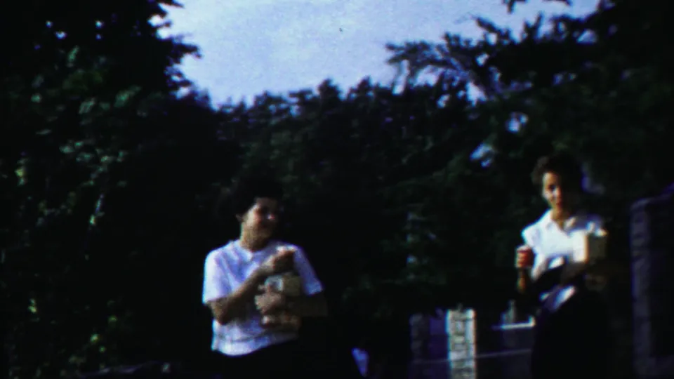 In the final frame, two young women remain near dense trees and masonry, one holding a light-colored item as the 8mm shot ends.