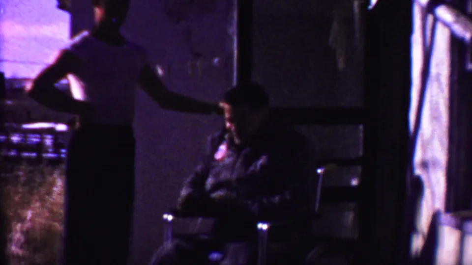 In the 1958 Kansas home-movie frame, a standing adult man gestures toward a seated adult man in a chair near a dark doorway.