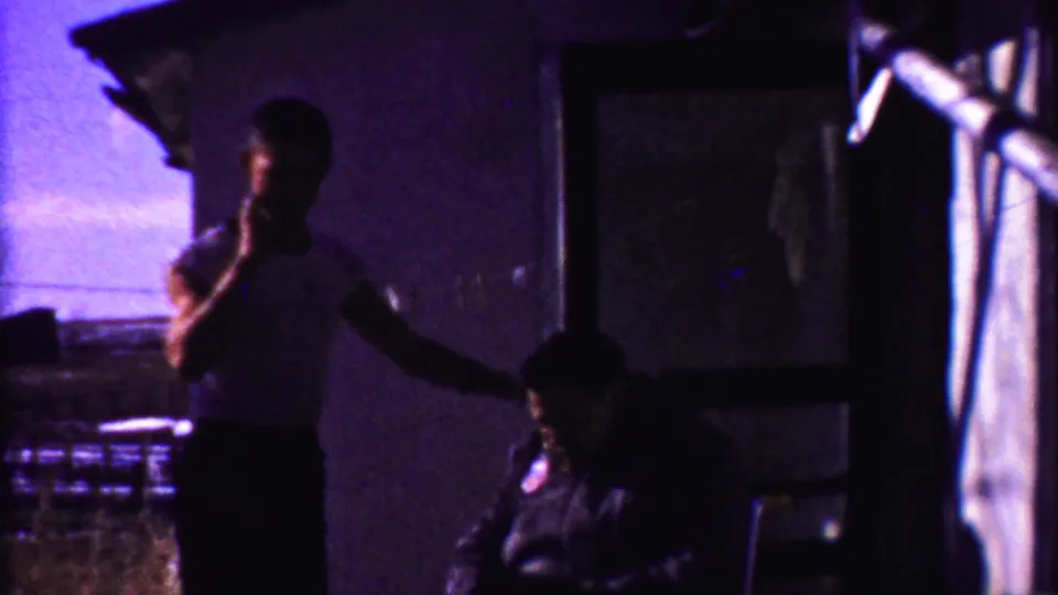 In this 1958 Kansas home-movie frame, a man in a white shirt appears to smoke while standing beside a seated man in porch shade.