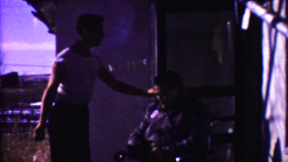 The son appears to reach toward or rest a hand near his seated father in a dim 1958 Kansas porch scene.