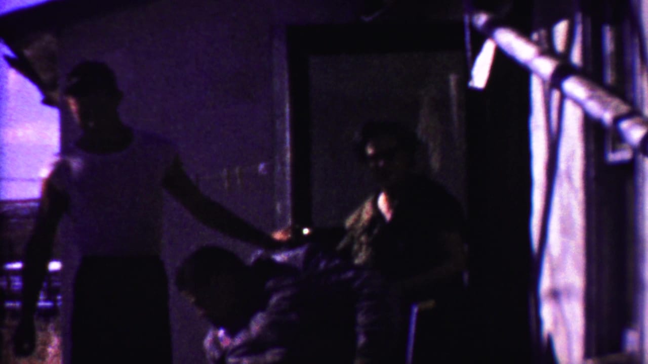 Three men outside a small building, with one standing near seated figures by a dark doorway.