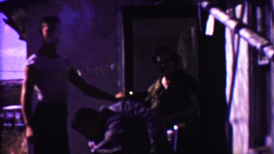 A man in a light shirt stands and gestures while two others remain near a doorway in rural Kansas home-movie footage.