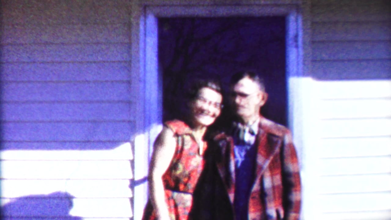 Adult couple standing in a house doorway, woman in red dress and man in plaid jacket.