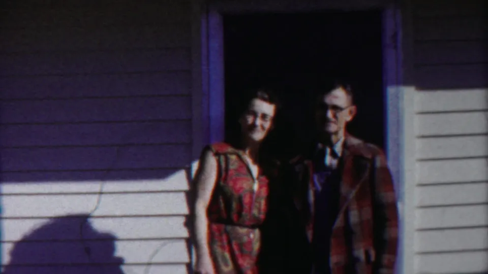 A 1958 Kansas home-movie frame shows the couple still posing, with the house wall and cast shadow prominent.