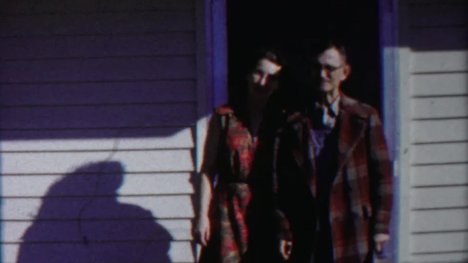 The couple remains in the doorway of a Kansas home, facing the camera as hard sunlight and shadow divide the siding.