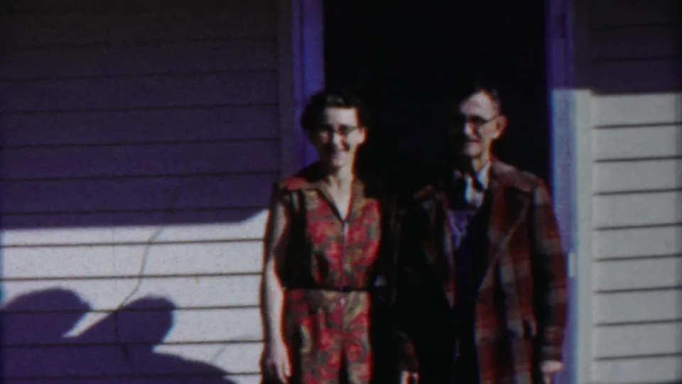 A woman in a red patterned dress and a man in a plaid jacket stand side by side in the doorway in this 1958 home movie frame.