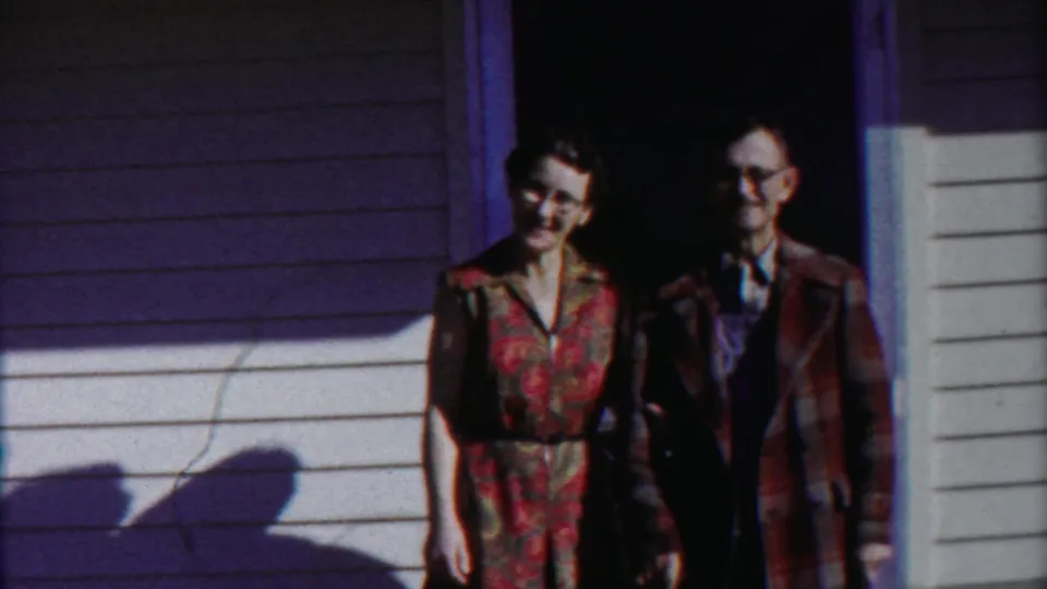 The couple remains near the doorway, with the woman's floral dress and the man's glasses visible in the 1958 Kansas home movie.