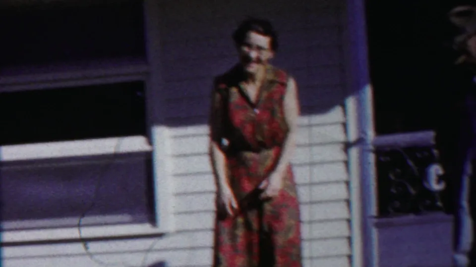 An adult woman in a floral dress stands alone near the house doorway and window in the Cheyenne County, Kansas home movie.