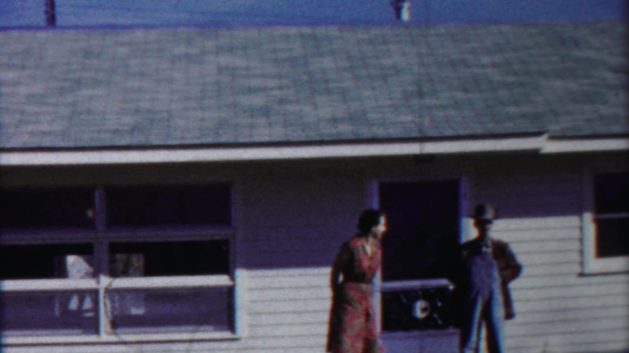 Two adults stand near a porch and window on a white-sided house.