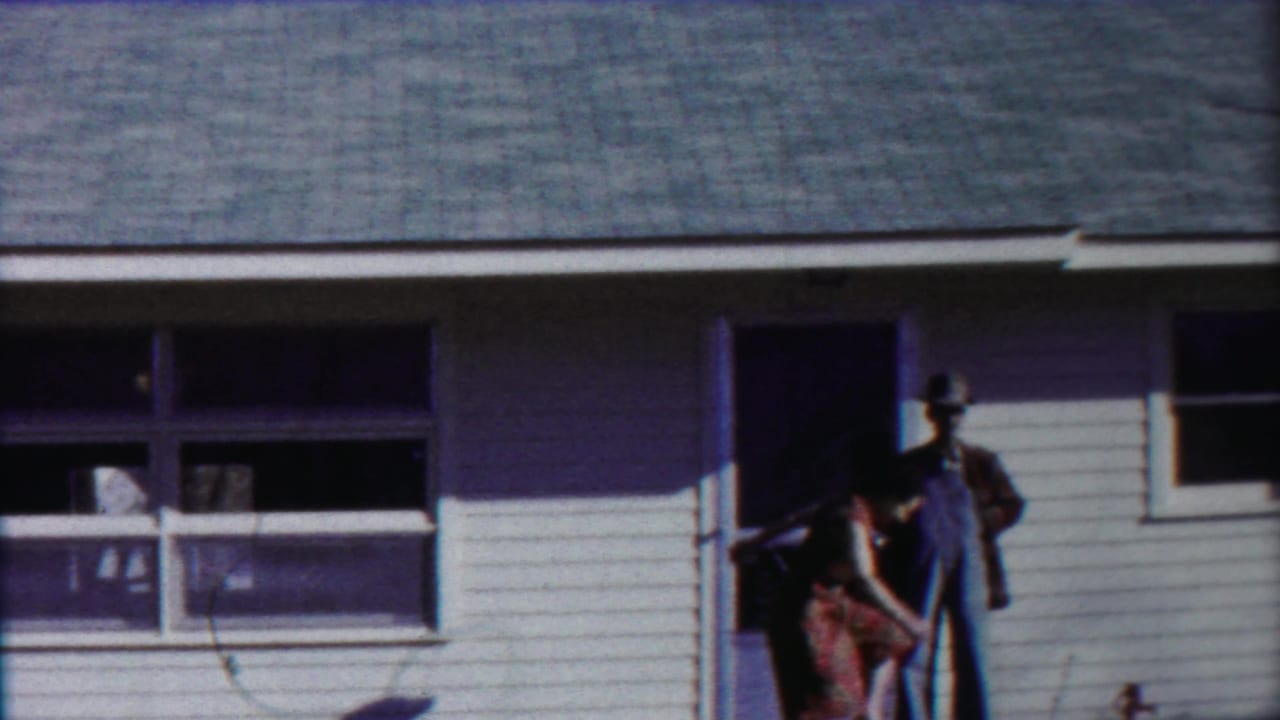 A man in overalls stands on a porch while a woman bends nearby.