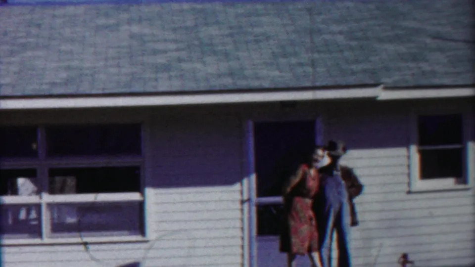 The woman in the red dress rises close to the man in overalls at the porch doorway in the 1958 Kansas home-movie scene.