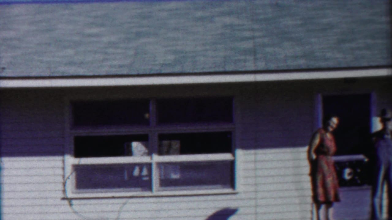 A white-sided house exterior shows windows, doorway edges, and partial figures near the right side.