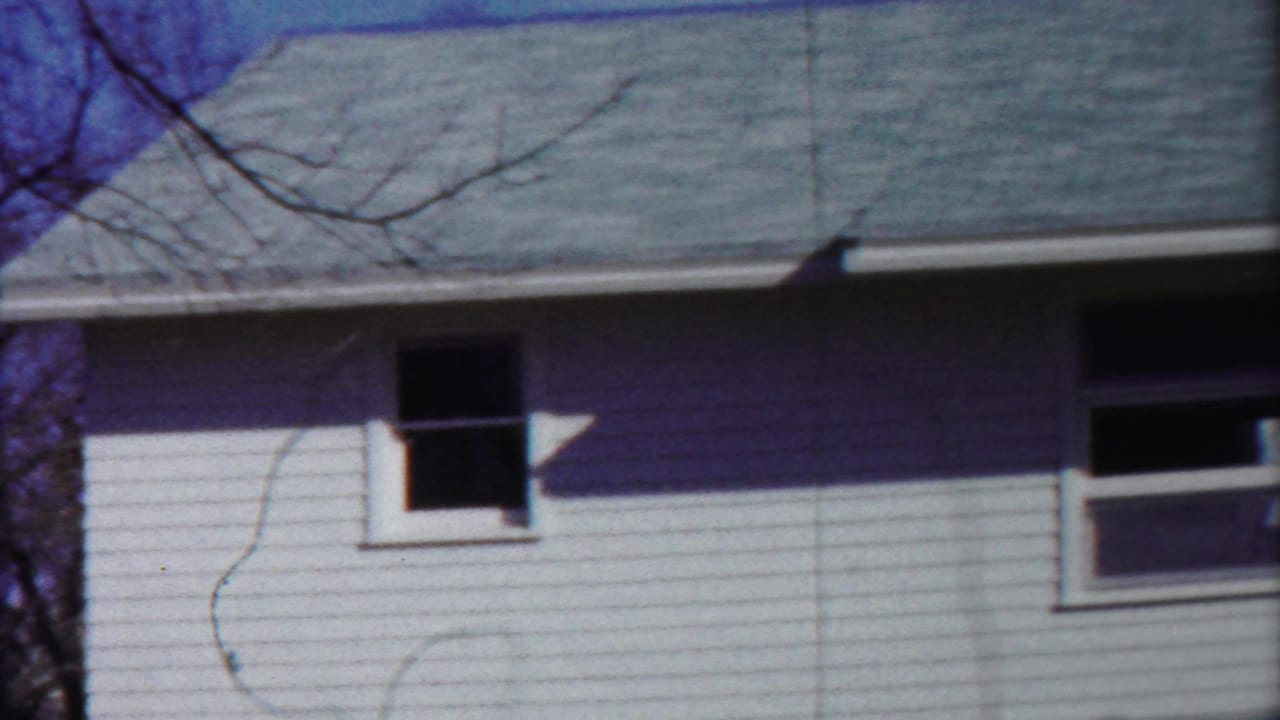 1958 Kansas home-movie frame showing a white-sided residential exterior with a gray roof, dark windows, and bare tree branches.