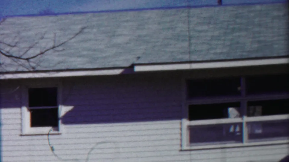 1958 Kansas frame of a white-sided house exterior with roof shingles, a small dark window, a larger front window, and bare branches.
