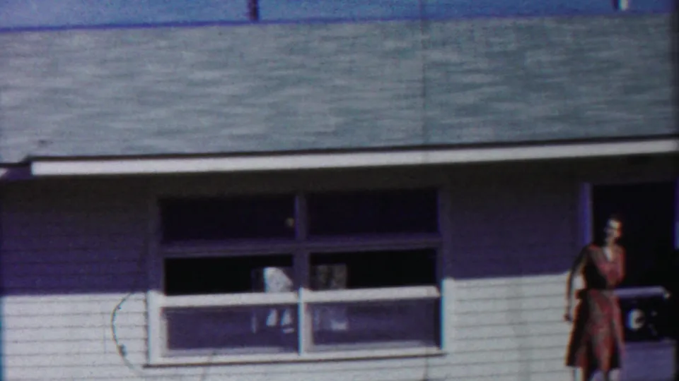 1958 Cheyenne County, Kansas frame showing a woman in a dress near the front doorway of a white-sided house, appearing to gesture outward.