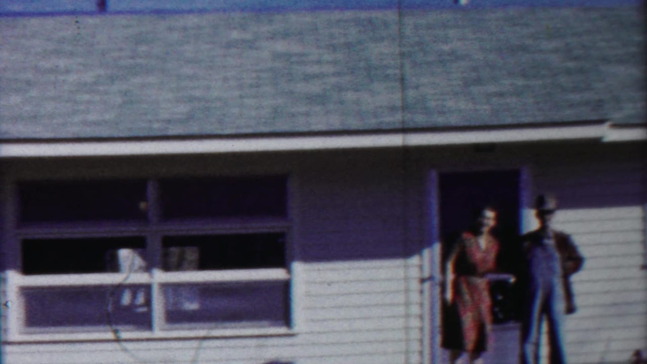 Woman in a dress and man in overalls stand by the open door of a white-sided house.