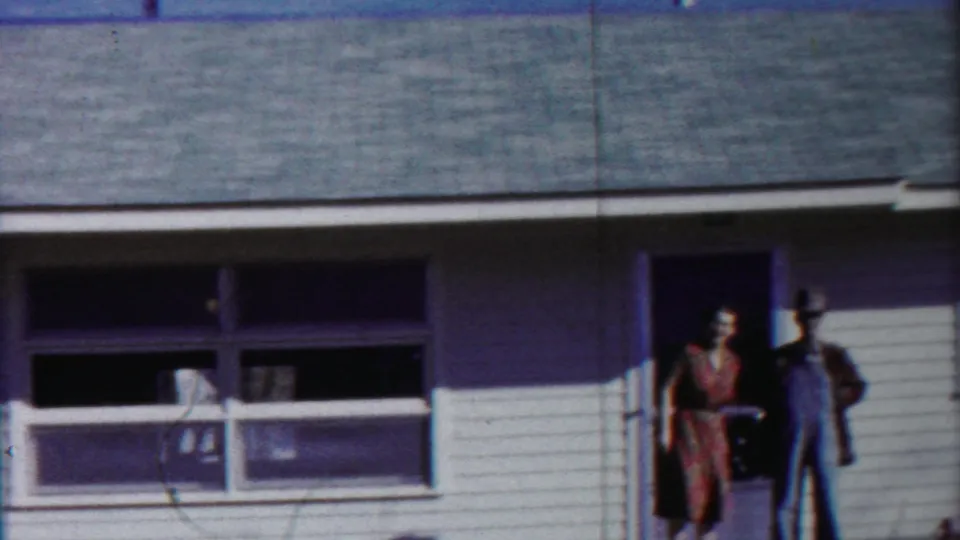 In supplied 1958 Cheyenne County footage, a woman in a patterned dress stands near a doorway beside a man in overalls and a hat.