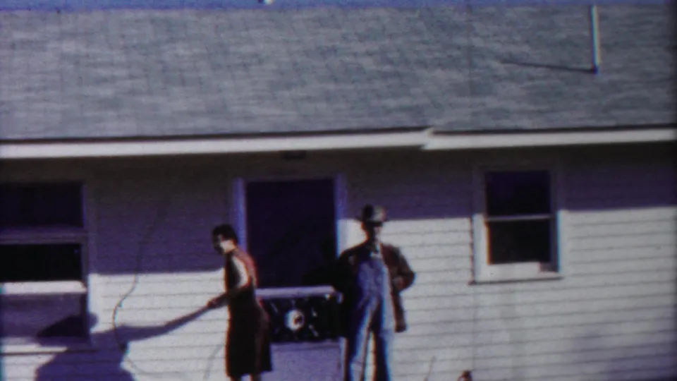 The woman moves across the front of the house while the man in overalls stays near the doorway in this rural Kansas home-movie frame.