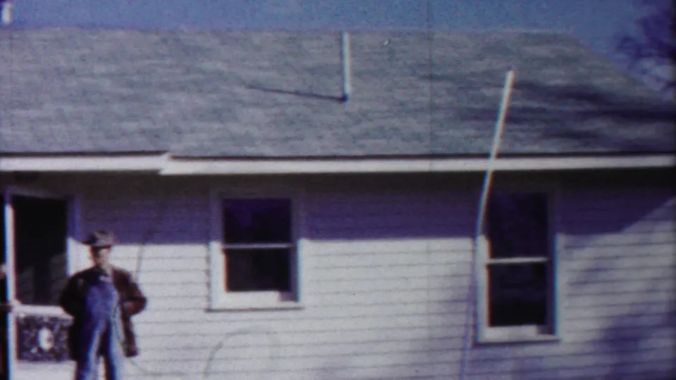 A man in overalls and a hat stands near the open door of a white-sided house in the supplied 1958 Cheyenne County home movie.