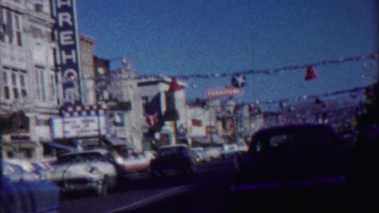 Street-level view of decorated storefronts, older cars, and hanging flags across the road.