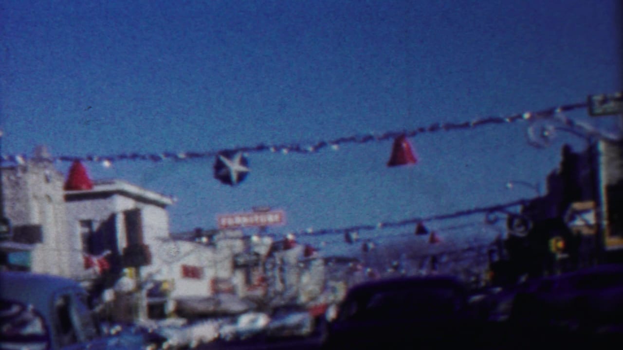 Decorated city street with overhead ornaments, storefronts, and cars under a blue sky.