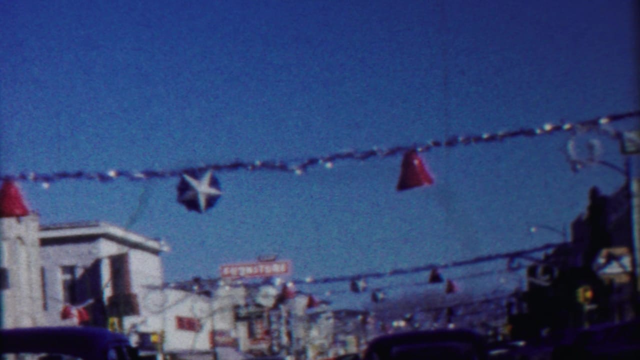 Moving 8mm view of a decorated Kansas commercial street, with star and cone ornaments suspended above cars and shopfronts.