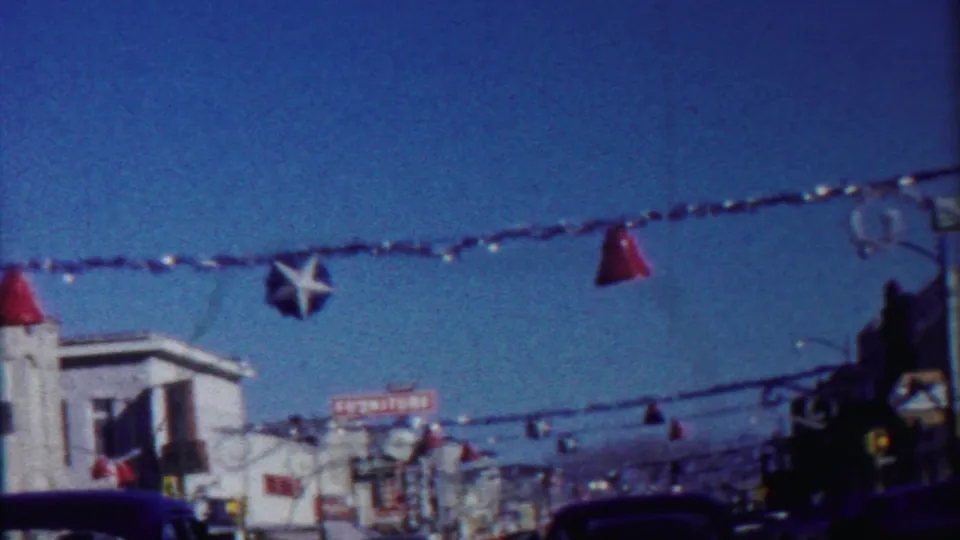 Moving 8mm view of a decorated Kansas commercial street, with star and cone ornaments suspended above cars and shopfronts.