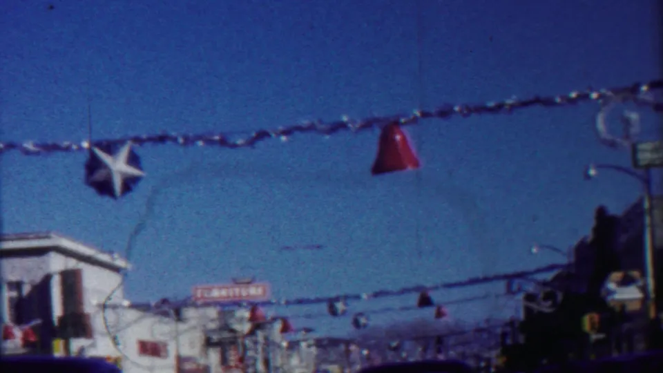 1950s home-movie frame showing a decorated Cheyenne County street with overhead garland, star ornaments, storefronts, and vehicles.