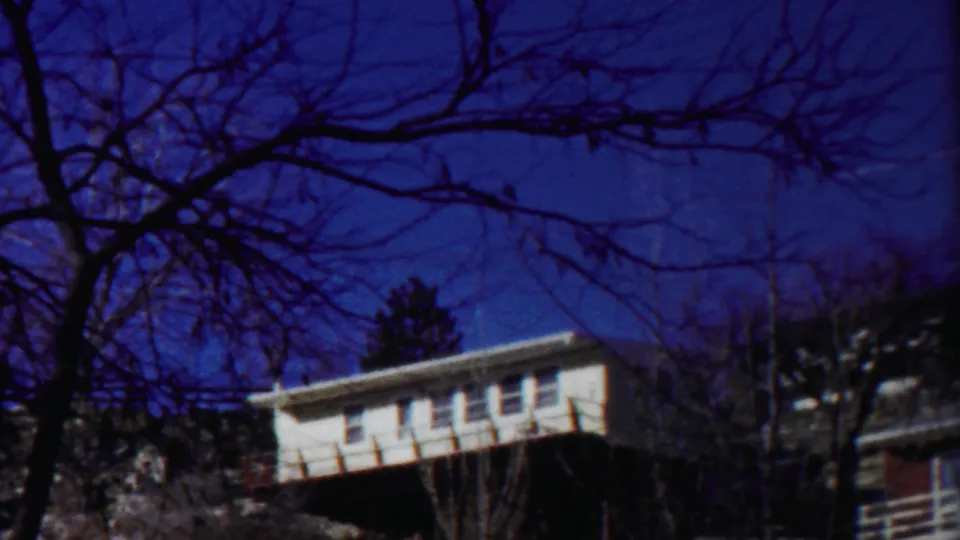 1958 Cheyenne County, Kansas 8mm frame showing a white house among bare trees on a hillside beneath a saturated blue sky.
