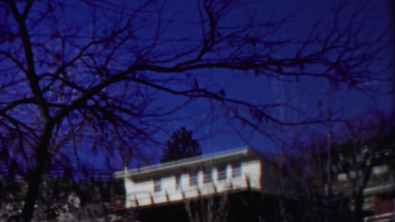 1958 Kansas home-movie frame with a white house, nearby darker structure, and winter-bare trees crossing the view.