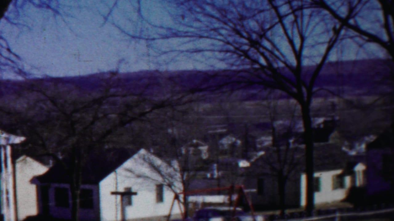 1958 home-movie frame of a Cheyenne County residential yard with modest houses, bare trees, a swing set, and a parked car partly visible.