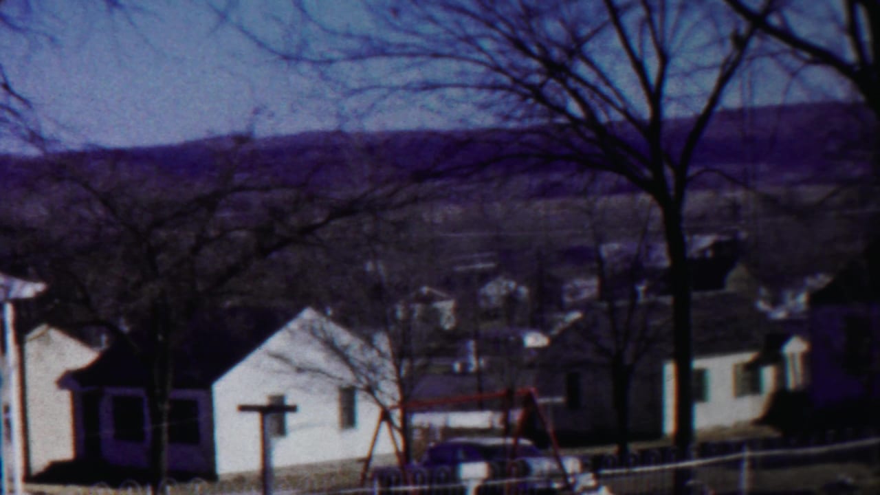 A soft 8mm frame shows a quiet 1958 Kansas residential street or yard with homes, swing set, parked car, bare trees, and distant hills.