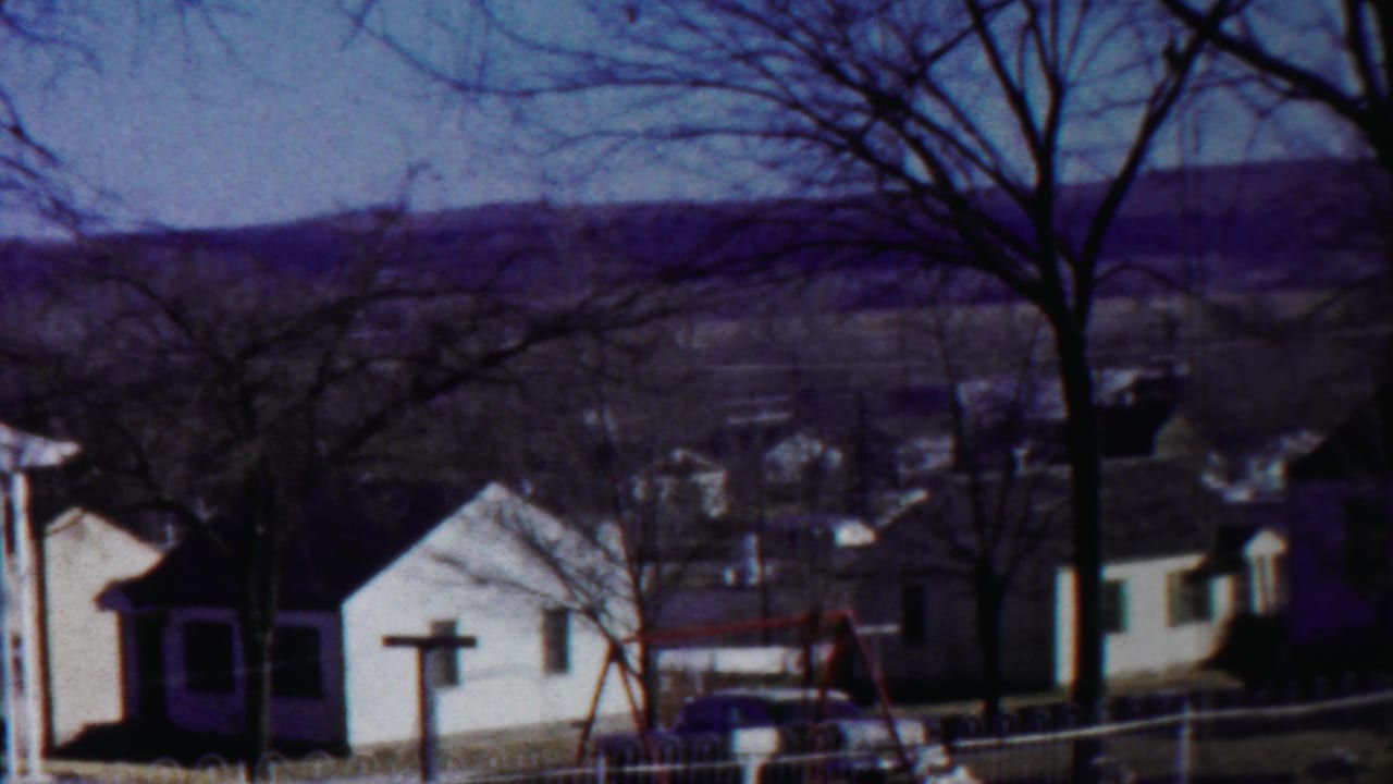 Residential street with white houses, bare trees, fence, swing set, and distant hills.