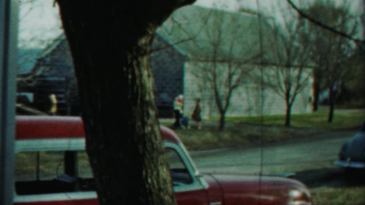 Home-movie frame showing a red mid-century vehicle, large tree trunk, residential road, modest homes, lawns, and distant people.