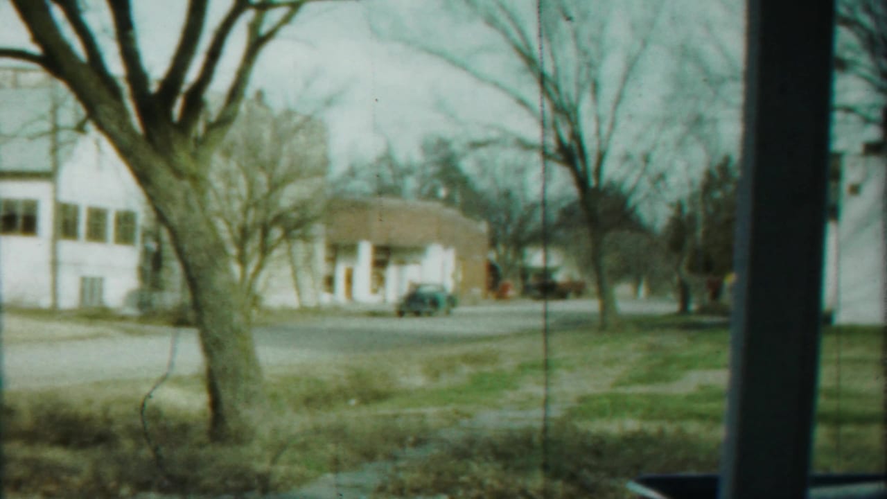 1958 Kansas residential street view with white houses, bare trees, parked car, open lawn, and small distant figures near the road.