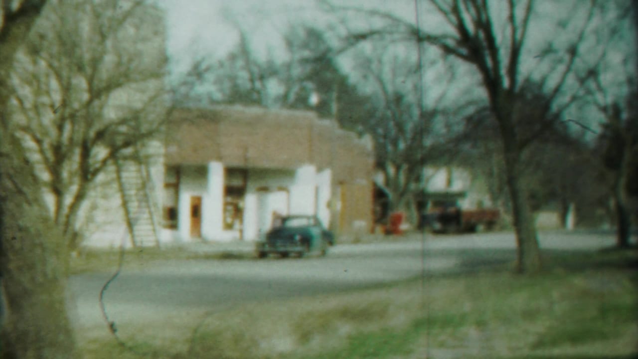 1958 Cheyenne County street scene with a modest storefront, parked dark sedan, bare trees, and a red truck in the background.
