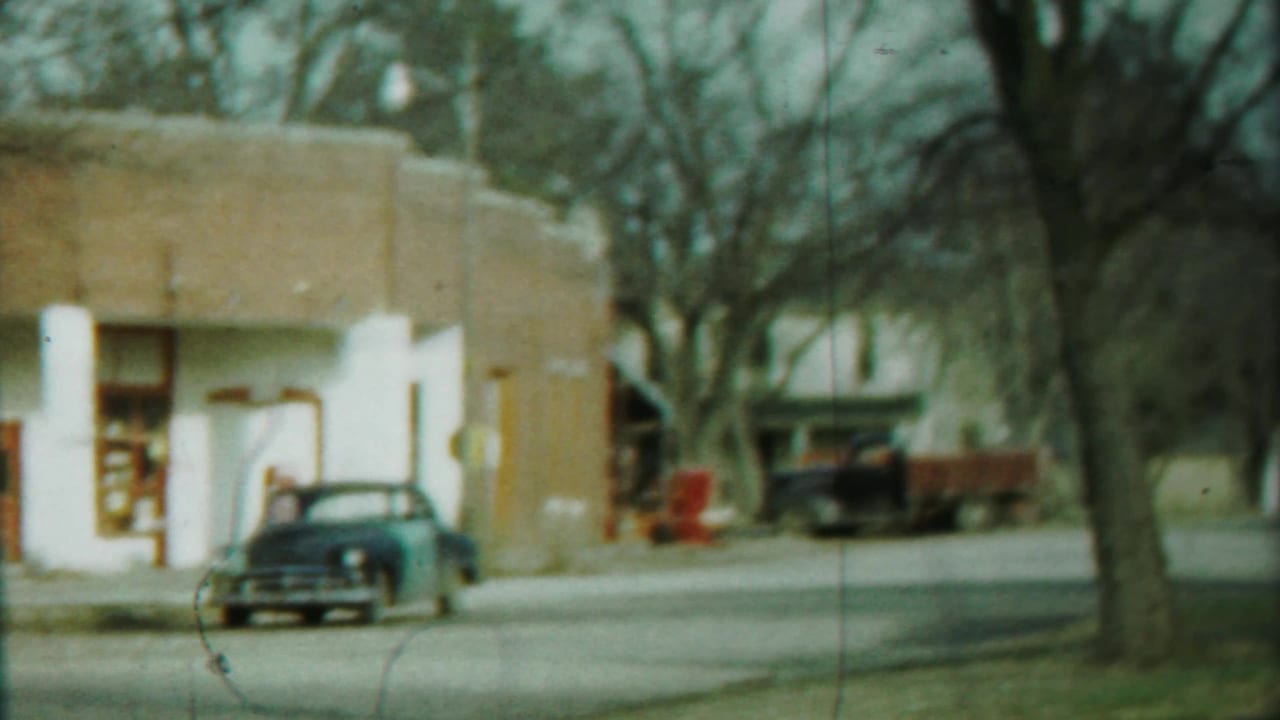 Soft 8mm view of a 1958 Cheyenne County roadside storefront, with a parked sedan, red truck, bare trees, and muted color.