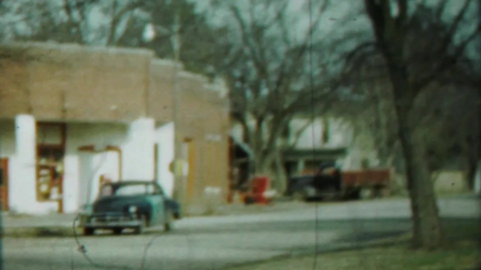 Soft 8mm view of a 1958 Cheyenne County roadside storefront, with a parked sedan, red truck, bare trees, and muted color.