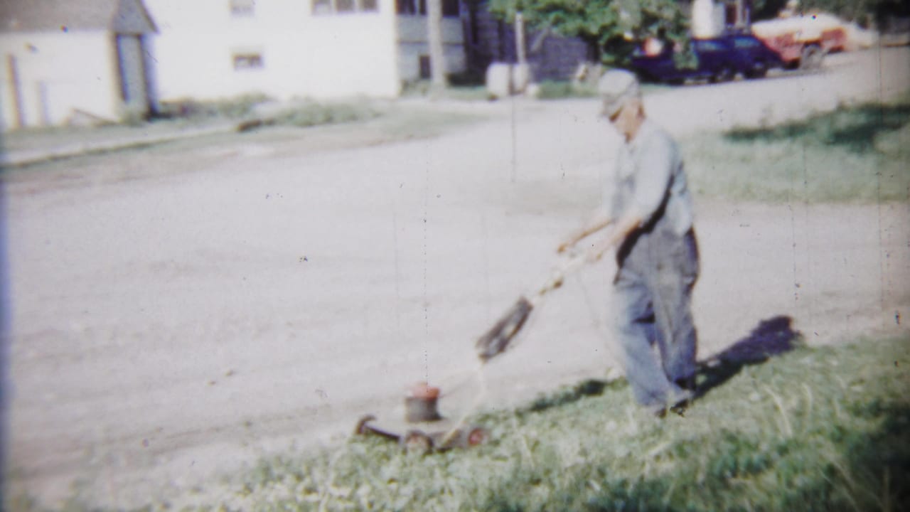Man in overalls pushes a lawnmower along grass beside a residential street.