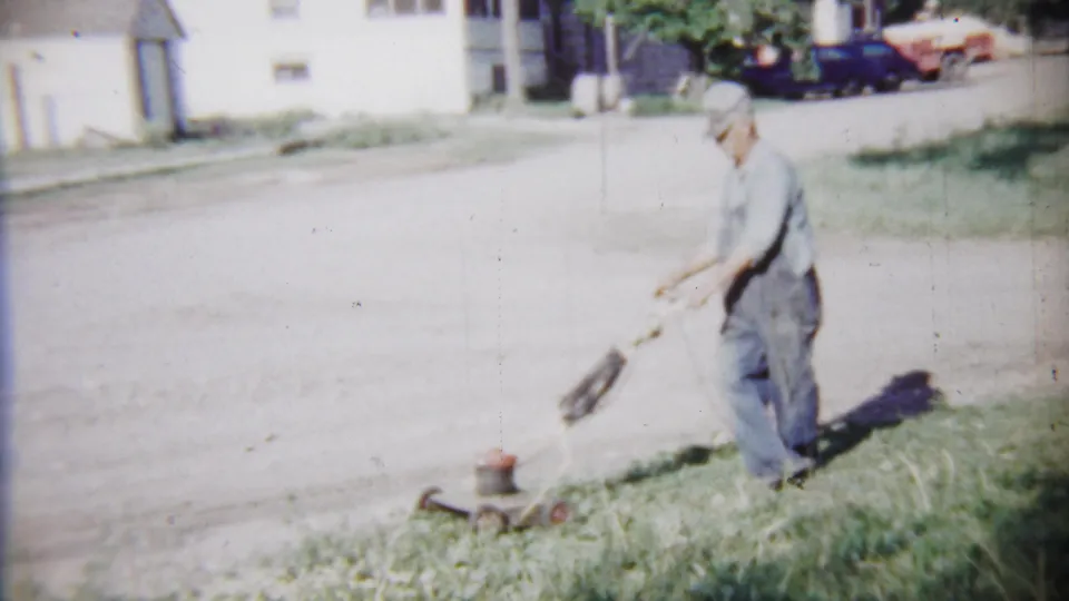 In 1958 Saint Francis, Kansas, an older man in overalls and cap mows a grassy edge near a quiet street and light-colored houses.